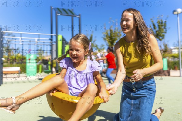 Mother and young daughter sharing laughter as mom pushes her on a bright yellow spinning cup at a colorful outdoor playground on a warm sunny day, carefree family fun