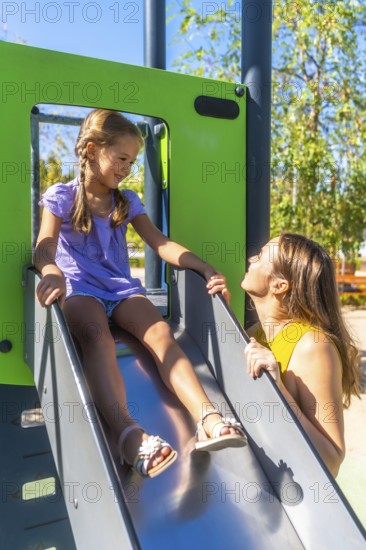 Mother encouraging her smiling daughter on a sunny park slide, sharing a playful bonding moment of support, joy and family connection during outdoor fun
