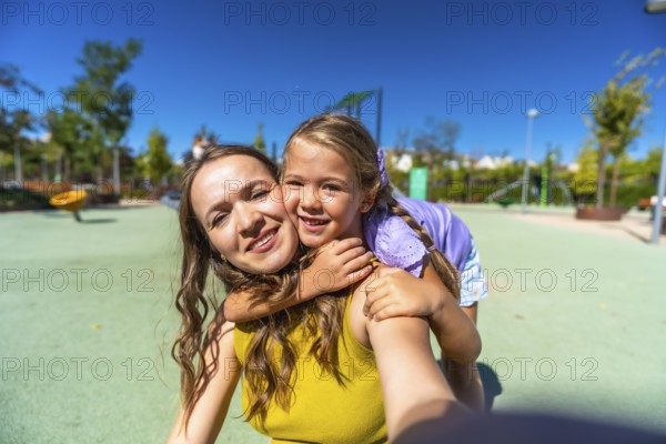 Mother and daughter smiling and embracing as they take a sunny playground selfie, enjoying a joyful day of family bonding, carefree laughter and warm summer fun