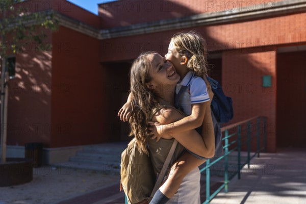 Mother holding her young daughter, both smiling and showing affection, standing outdoors near a school building with backpacks, representing family support and the back to school experience