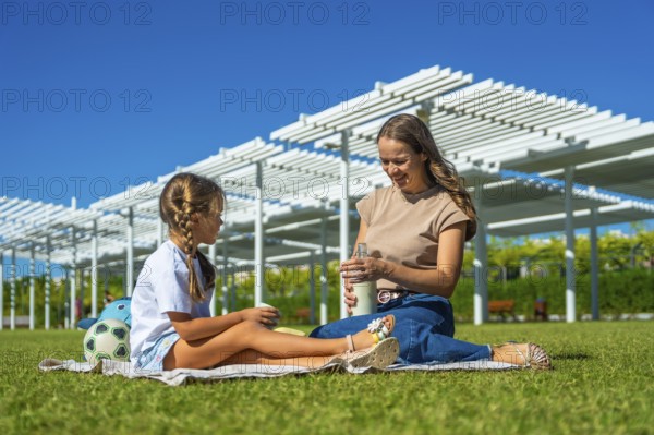 Mother and daughter sitting on a picnic blanket in a sunny urban park, smiling and bonding over a bottle of milk during a relaxed summer outing among green grass and blue sky