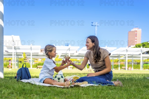 Mother and daughter spending quality time together on a picnic blanket in a sunny park, sharing a snack and smiling, highlighting family bond and happy summer moments