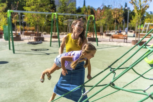 Mother and daughter smiling and laughing while playing on a sunny outdoor playground, climbing rope net and enjoying warm daytime family bonding in a green urban park