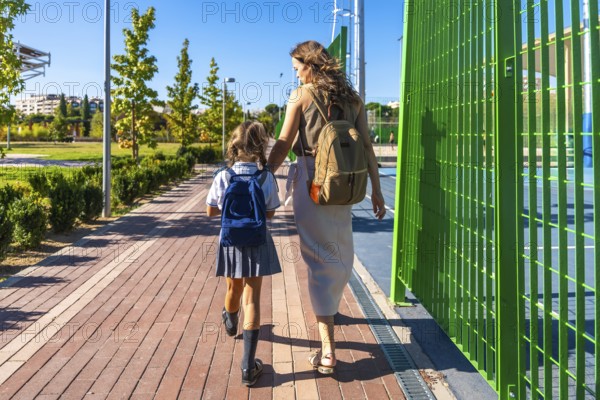 Mother and daughter walk hand in hand on a sunny sidewalk, child in school uniform and backpack heading to class as mom offers support and encouragement on the first day back