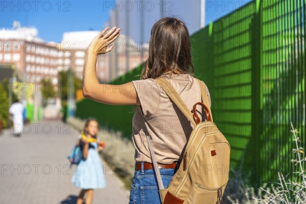 Mother waving goodbye, watching her young daughter with a backpack walk independently towards school, symbolizing back to school, parenting, and family separation emotions
