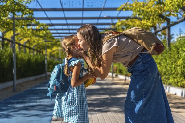 Woman and little girl sharing an affectionate moment while going to school, mother giving her daughter a kiss on the cheek, celebrating family love and education