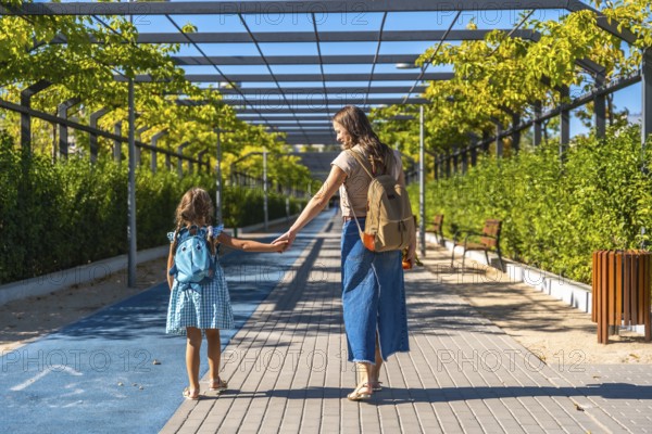 Mother and daughter walking along an urban path, holding hands and carrying backpacks on a sunny day. This image represents themes of family, education, and childhood