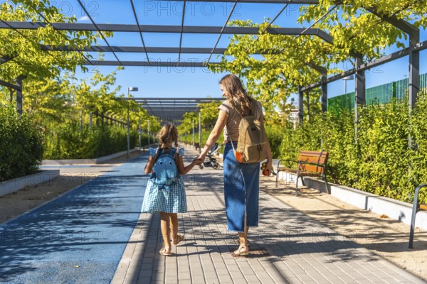 Mother and daughter, wearing backpacks, walking along a sunny pathway lined with green trees and bushes, holding hands on their way to school or kindergarten