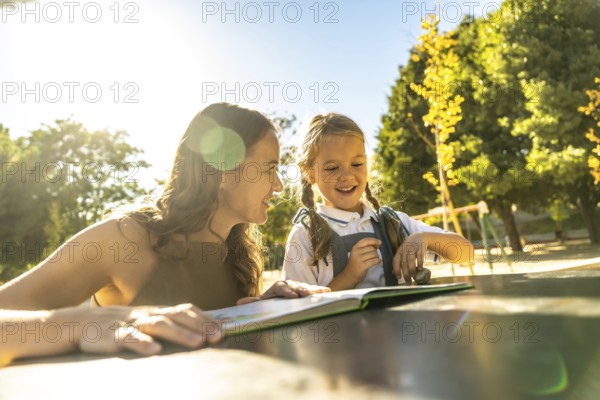 Mother and daughter smiling and laughing while reading a book together at an outdoor table in a sunny park or school playground, enjoying warm quality time and learning