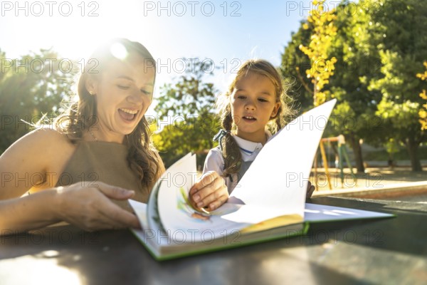 Mother and daughter engaging in a fun reading activity, turning pages of a colorful book while learning and laughing together outdoors during a sunny school day