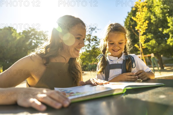 Mother and daughter smiling together at an outdoor schoolyard table, reading a book and bonding over learning in sunshine, casual uniforms and leafy surroundings