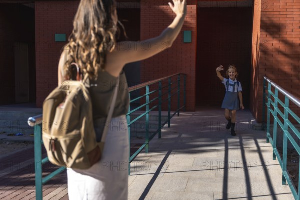 Mother and daughter saying goodbye, the child waving to her mother while walking into school wearing a uniform and backpack, symbolizing back to school and education