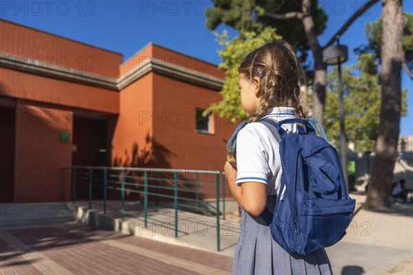 Young schoolgirl wearing a uniform and carrying a blue backpack, walking towards the entrance of an elementary school building on a sunny day, preparing for her education