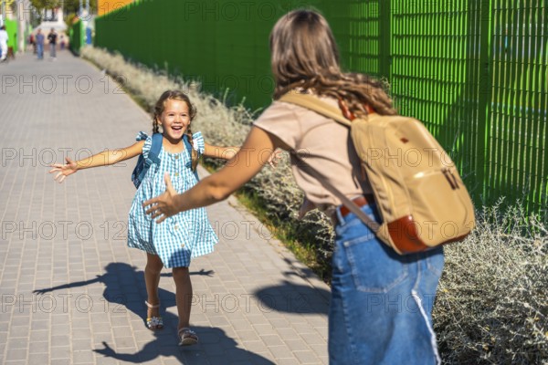 Child daughter with backpack runs across schoolyard to joyfully hug her mother at pickup, celebrating a warm reunion of family love and connection after a school day