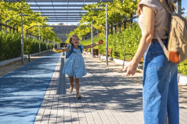 Excited schoolgirl with backpack runs toward her mother in a sunny park, holding a banana after school, joyful reunion and carefree summer energy between parent and child
