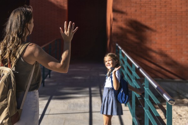 Mother waving goodbye to her elementary school daughter holding a backpack and happily walking towards the school building, starting a new day of education