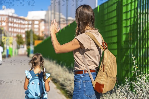 Mother waving with her hand to a small daughter carrying a backpack and walking away towards school, symbolizing childhood independence, family support, and the back to school concept