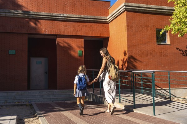 Mother holding hands with her daughter, both walking together towards a brick school building on a sunny day, representing family support and the start of education
