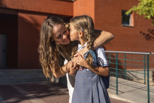 Mother embraces and kisses her young daughter in school uniform at the school entrance on the first day, tender back to school moment filled with love and support outdoors