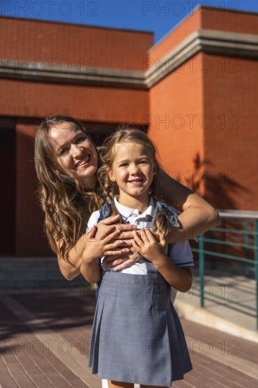 Mother and daughter smiling while standing together on a sunny day at school with the elementary schoolgirl wearing her uniform and backpack, symbolizing support and care