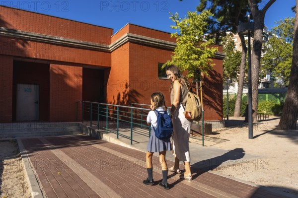 Mother and daughter smiling as they walk toward a red brick school building with backpacks, enjoying a sunny back to school morning and bonding before the new academic year