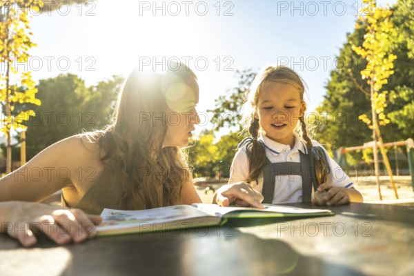 Mother and daughter bonding over reading a book together, sitting at a table outdoors in a bright sunny school environment, focusing on education and learning