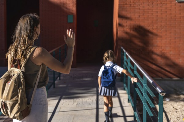 Mother waving goodbye to her elementary age daughter wearing a school uniform and backpack, walking inside a brick school building on a sunny day during drop off