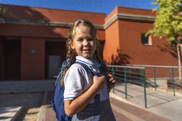 Young female student smiling and looking at the camera, wearing a school uniform and carrying a backpack, standing outdoors at a school campus on a sunny day