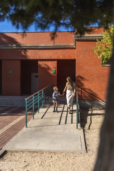Mother and daughter holding hands and walking together, approaching a school building on a sunny day, symbolizing back to school, education, and family support