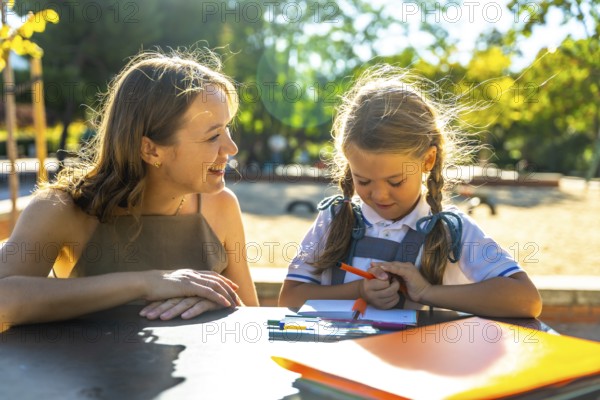 Mother and daughter sitting at a table outdoors in a park, engaging in a school activity and drawing together, fostering a positive learning environment