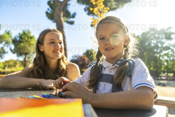 Young daughter and her mother enjoying a sunny day outdoors at the school playground, the child drawing on a table, representing education, family, and childhood