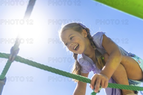 Happy young girl laughing with joy while actively climbing a green rope net structure outside on a sunny day, experiencing fun during childhood playtime