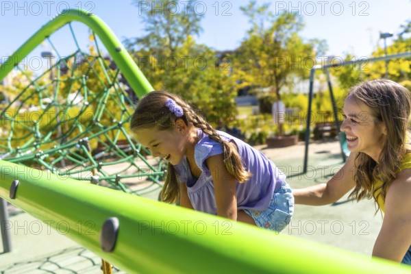 Playful daughter laughing as she climbs a green playground structure while mother smiles and supports her, sharing a joyful outdoor moment of family bonding and carefree summer fun