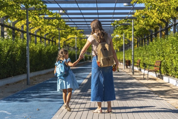 Mother and daughter holding hands and walking together along a paved path, carrying backpacks and heading towards school on a bright sunny day, ready for learning