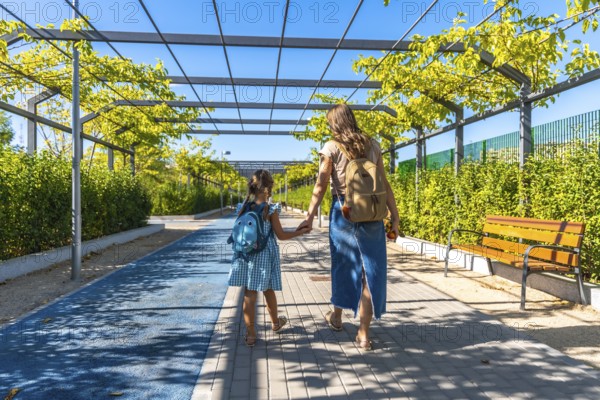 Mother and daughter walk hand in hand along an urban park path with backpacks, heading to school on a sunny daybonding, guidance, and a fresh start toward learning and growth