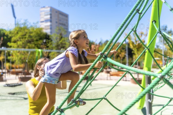 Mother assisting her happy daughter climbing a green rope jungle gym on a sunny day, experiencing childhood fun and family activity together in a park