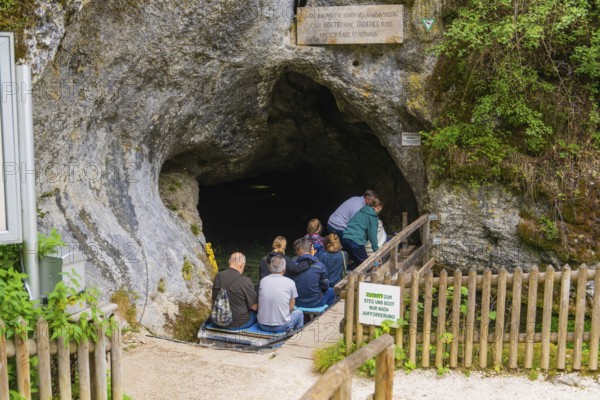 Group of people at the entrance of a cave surrounded by rock walls and lush vegetation, Wimsener Höhle, Hayingen, Germany