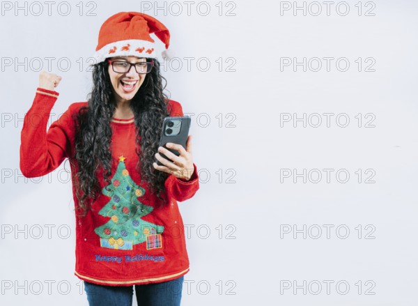 Excited girl in Christmas sweater looking at phone screen, isolated. Young woman in Christmas hat celebrating and looking phone