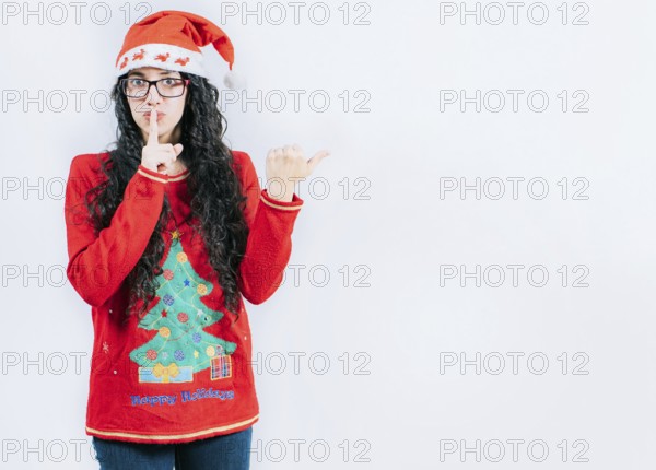 Girl in Christmas sweater and glasses pointing to the side, isolated. Young woman in Christmas hat making secret gesture recommending an advertisement