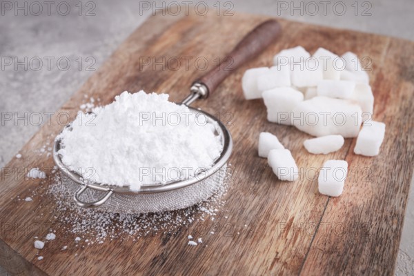 A strainer with powdered sugar, on a wooden cutting board, with pieces of white sugar