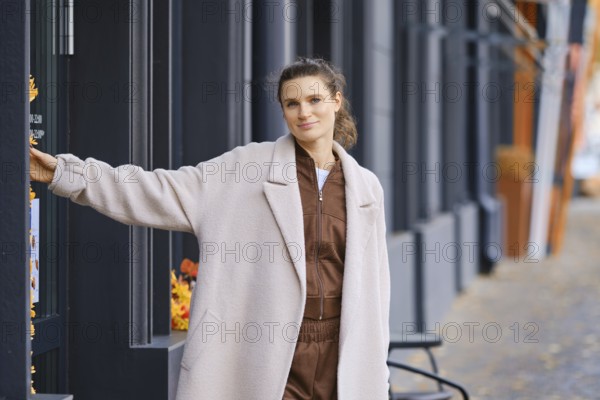 A fashionable woman is posing near a cafe entrance in a modern urban area. She wears a cozy outfit and smiles while leaning against the door, enjoying the pleasant atmosphere of the day