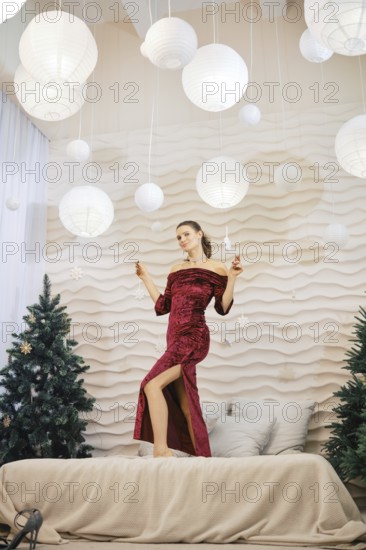 A stylish woman in a red dress stands playfully on a bed, surrounded by soft lights and Christmas trees