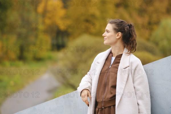 A woman stands calmly in an autumn landscape, surrounded by golden trees. She wears a cozy outfit and smiles while gazing thoughtfully into the distance, embodying serenity in nature