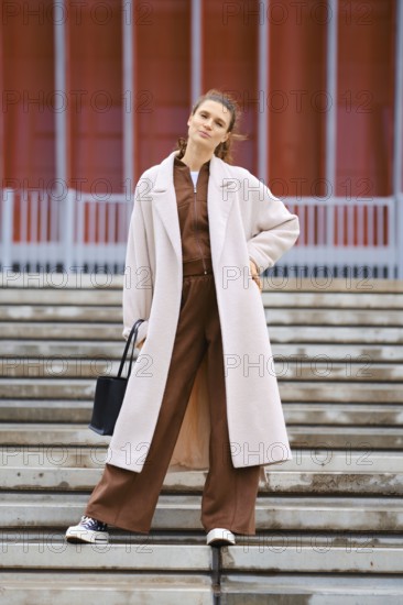 A confident woman stands on the steps wearing a brown tracksuit and a long cream coat. She carries a black bag and smiles, presenting a fashionable look against a contemporary background