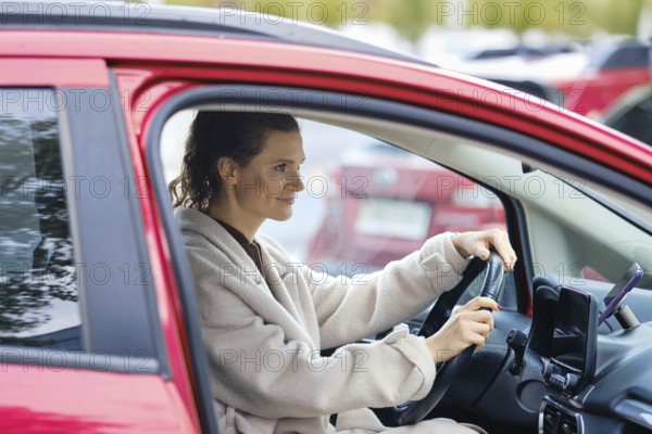 A woman sits in the driver's seat of a red car in a busy parking lot. She has a confident expression as she holds the steering wheel, preparing to drive
