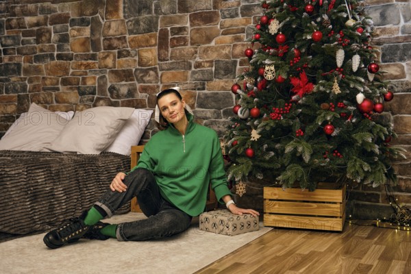A cheerful woman sits on the floor beside a beautifully decorated Christmas tree in a cozy indoor space. The room features rustic stone walls and festive decorations that create a warm atmosphere