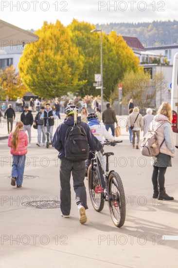 A man pushes his bike across a sidewalk while people and autumn trees form the backdrop, Urschelherbst Street Festival, Nagold, Germany
