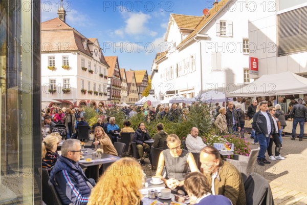 People sit in a street café in an old town under sunny skies. Busyness and convivial atmosphere prevail, Urschelherbst street festival, Nagold, Germany