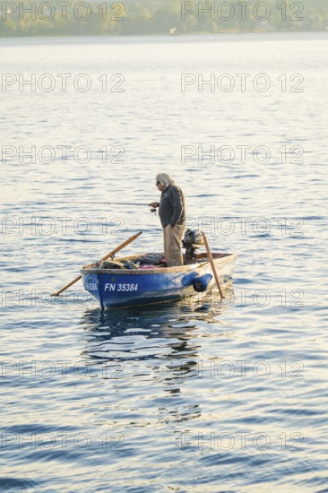 A man stands in a small blue boat fishing in a peaceful lake, Überlingen, Lake Constance, Germany