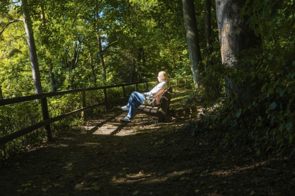 Woman sitting relaxed on a bench in the forest, pierced by sunspots, Überlingen, Lake Constance, Germany